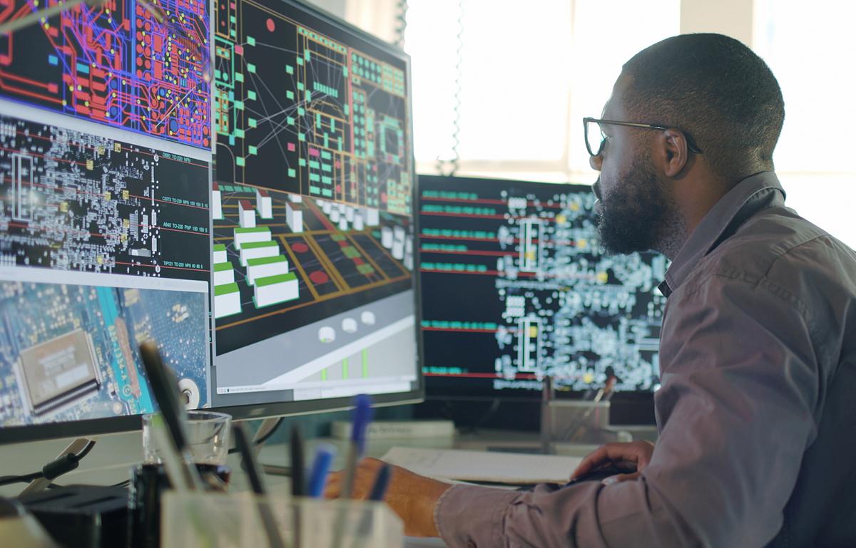 Stock image of an Afro-Caribbean male designing electronic circuit boards ( PCBs). He’s sitting at a desk with a large computer screen displaying a variety of apps & PCB illustrations.